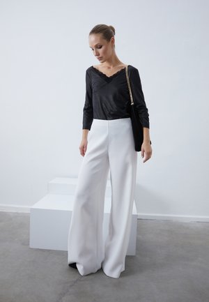 Μαύρη μπλούζα με δαντελένια λεπτομέρεια, συνδυασμένη με φαρδιά λευκά παντελόνια. Το μοντέλο κρατά μια μαύρη τσάντα με αλυσίδα. Απλός, μοντέρνος σχεδιασμός και χρώματα.