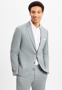 Hellgrauer Anzug aus strukturiertem Stoff, mit einem Zweiknopf-Blazer, taillierter Passform und kontrastierenden braunen Knöpfen. Weißes Hemd darunter.