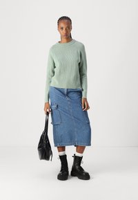 Minzgrüner gerippter Pullover, Jeans-Midi-Rock mit seitlichen Taschen, schwarze Schnürstiefel und eine schwarze Handtasche. Verstellbare Passform mit ausgeprägten Texturen.