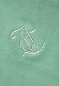 Tessuto verde menta con una superficie liscia presenta iniziali ricamate in bianco, mostrando intricati anelli e curve del design.