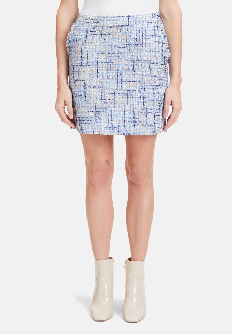 Femme portant une mini-jupe à carreaux bleu et beige texturée et des bottines crème en cuir verni, les mains détendues le long du corps.