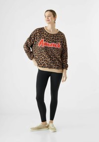 Lepaardprint sweatshirt met rode "Amour" tekst, lange mouwen, ribbelzoom, gecombineerd met zwarte leggings en beige sneakers. Zachte stofstructuur.