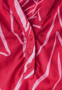 Rotes Stoffoberteil mit einem pinken Zickzackmuster, V-Ausschnitt und vier passenden Knöpfen auf der Vorderseite. Glatte Textur mit sichtbaren Nähten.