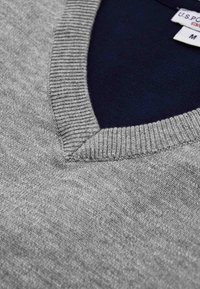 Grauer V-Ausschnitt-Pullover mit geripptem Kragen, zeigt ein dunkelblaues Innenfutter und ein teilweise sichtbares weißes Kleidungsetikett in Größe M.