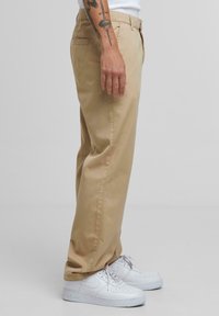 Beige bukser laget av bomull, med rett ben design, sidelommer og en glatt tekstur. Båret med hvite sneakers.