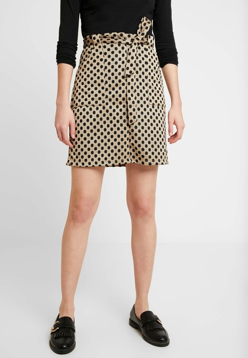 Femme portant une jupe beige à pois noirs, une ceinture nouée, des poches avant, un haut noir à manches longues et des mocassins noirs.