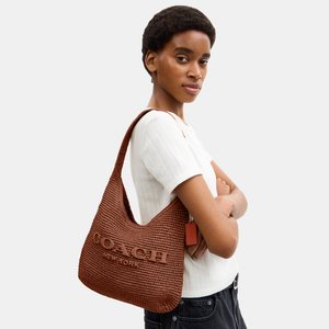 Giovane donna con camicia bianca e jeans neri porta una grande borsa a tracolla marrone intrecciata Coach New York sulla spalla.