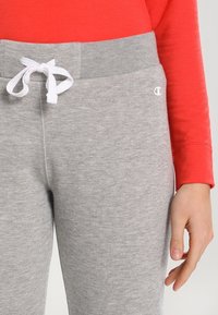 Graue Sweatpants mit einer weichen Textur, ausgestattet mit einem weißen Kordelzug und einem Logo auf dem linken Oberschenkel, kombiniert mit einem leuchtend roten Langarmshirt.