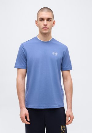 Ung mand med kortklippet hår iført en blå T-shirt med EA7-logo og sorte bukser med gyldent tryk, stående foran en ensfarvet lys baggrund.