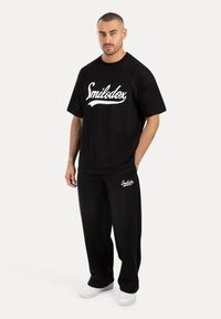 Czarny bawełniany t-shirt z białym logo "Smilodox", w połączeniu z czarnymi dresami z dopasowanym logo, dopełniony białymi sneakersami.