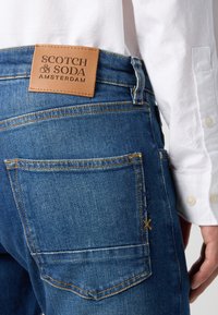 Blaue Denim-Jeans mit einem tanfarbenen Leder-Patch mit "SCOTCH & SODA" Branding, sichtbaren Steppdetails und einem Rücktaschendesign.