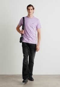 Camiseta corta de manga en color lila con un pequeño logo negro, combinada con pantalones negros y zapatos negros, llevada con un bolso cruzado negro.
