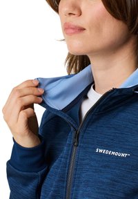 Marineblaues Zip-Jacket aus strukturiertem Stoff; hellblauer Innenkragen; mit dem Logo "SWEDEMOUNT" auf der Brust; Hand hält den Kragen.
