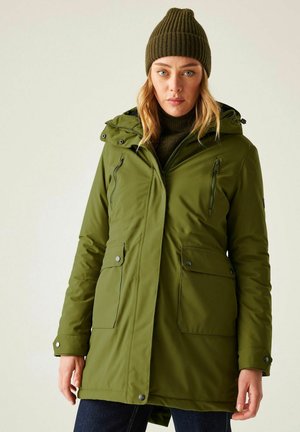 Vrouw draagt een olijfgroene winterjas met capuchon over een donkergroene trui en bijpassende gebreide muts, staand tegen een effen lichte achtergrond.