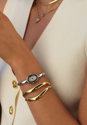 Arm mit silberner ovaler Uhr und zwei goldenen, wellenförmigen Armreifen, bekleidet mit weißem, strukturiertem ärmellosem Kleidungsstück mit goldenen Knöpfen und geschichteten Halsketten.