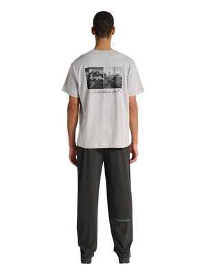 Man draagt een grijs T-shirt met zwart-witte landschapsfoto's en een zwarte broek met groene cijfers op het linkerbeen, staand met de rug naar ons toe.