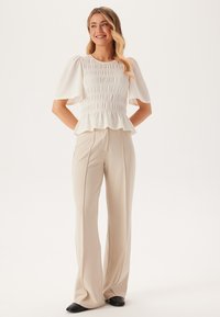 Top bianco a balze con maniche corte a sbuffo, abbinato a pantaloni beige a vita alta e scarpe nere a punta. Tessuto liscio e vestibilità comoda.