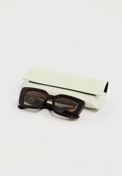 Ορθογώνια μαύρα γυαλιά ηλίου με σκούρους φακούς τοποθετημένα πάνω σε θήκη από ανοιχτό μπεζ δέρμα σε λευκό φόντο.