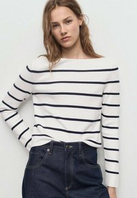 Witte blouse met lange mouwen en marineblauwe horizontale strepen, met een bootnek. Gecombineerd met hoge donkere spijkerbroeken.