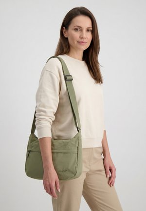 Kvinde iført cremefarvet sweatshirt og beige bukser bærer grøn skuldertaske med justerbar rem, stående mod en ensfarvet baggrund.