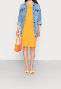 Żółta sukienka bodycon z dżinsową kurtką, pomarańczową torebką i jasnoniebieskimi klapkami. Dżinsowa kurtka zapinana na guziki; torebka ma wzór krokodyla.