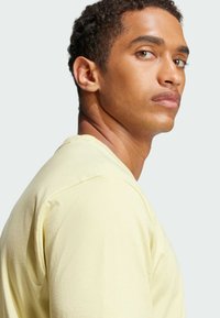 Světle žluté tričko s krátkým rukávem, kulatým výstřihem a měkkou tkaninou. Model směřující vlevo, ukazující jemnou pleť a strukturované vlasy.