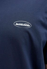 Sötétkék pamut póló, fehér ovális logóval, rajta a "Jack & Jones" és a "Club 1990" szöveggel, rövid ujjú, klasszikus kerek nyakkivágással.