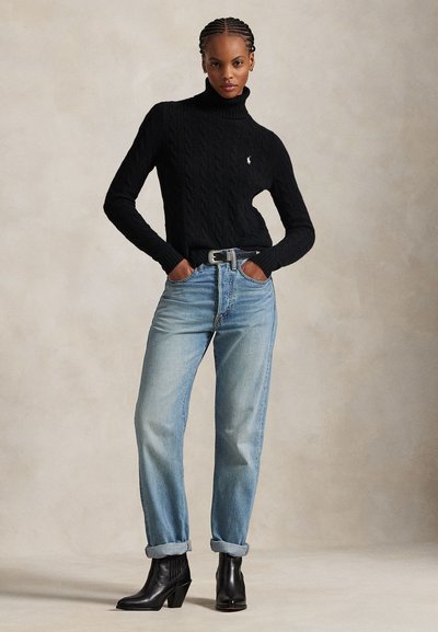 Donna con capelli intrecciati che indossa un maglione a collo alto nero, jeans azzurri risvoltati, cintura nera e stivaletti neri, in piedi con le mani nelle tasche.