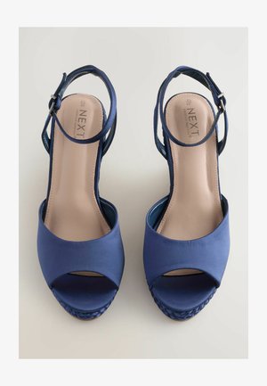 Tamno plave sandale s visokim potpeticama, otvorenim prstima i remenom oko gležnja, koje imaju pletenu platformu. Vidljiv logo branda unutra.