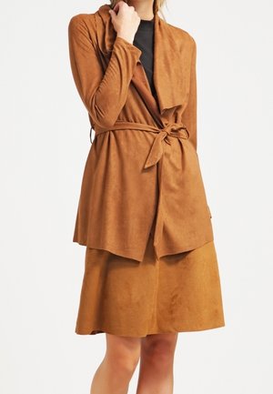 Femme portant un manteau en daim marron ceinturé à la taille sur une jupe assortie longueur genoux et un haut noir.