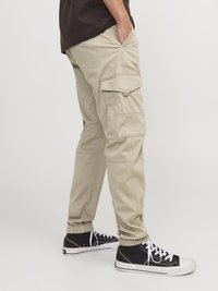 Pantaloni cargo bej cu buzunare laterale, picioare conice și manșete elastice. Asortați cu adidași negri cu glezna înaltă și șireturi albe.