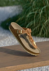 Rudos Birkenstock sandalai su sagtimi ir pintu dirželiu, gulintys ant medinės paviršiaus, aplink kuriuos žalia veja ir išblukusi fono. 