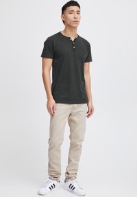 Camisa henley de manga corta gris oscuro con tres botones, combinada con pantalones beige y zapatillas blancas con rayas negras. Diseño simple y ajuste cómodo.