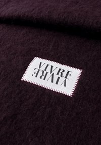 Tissu texturé violet foncé avec une étiquette rectangulaire blanche cousue en fil bordeaux affichant le texte inversé "VIVRE."