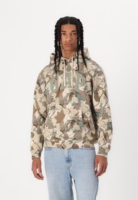 Beige huvtröja med ett camouflagemönster av blad i grönt och brunt, med "GAP" tryckt i stora bokstäver över bröstet.