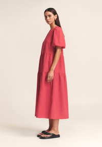 Robe rouge à étages avec manches courtes bouffantes et encolure ronde. Fabriquée en tissu lisse, tombant juste au-dessous de la cheville, portée avec des sandales noires.