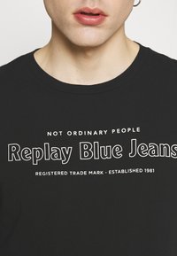Mustat puuvillapaidat, joissa on pyöreä pääntie, ja valkoiset tekstit: "EI TAVALLISET IHMISET" ja "Replay Blue Jeans" tavaramerkkitiedot alla.