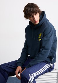 Mörkblå hoodie med en känguruficka och en gul grafisk design på bröstet. Tyget verkar slätt och mjukt; kombinerad med mörkblå byxor.