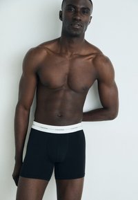 Boxer in cotone nero con una vita bianca su cui è scritto "Calvin Klein". Il tessuto appare liscio ed elastico. Il modello ha un torso ben definito.