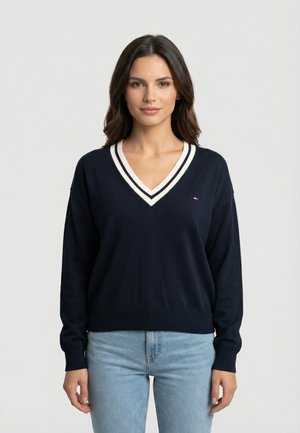 Femme aux cheveux foncés portant un pull bleu marine à col en V avec des rayures blanches et un jean bleu clair, debout devant un fond clair uni.