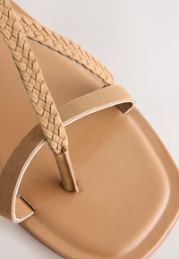 Brun flad sandal med flettede remme, en glat fodseng og et minimalistisk design. Sålen er afrundet med en ren finish.