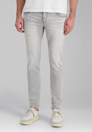 Mann trägt hellgraue Slim-Fit-Jeans und weiße Sneaker und steht vor einem schlichten grauen Hintergrund.