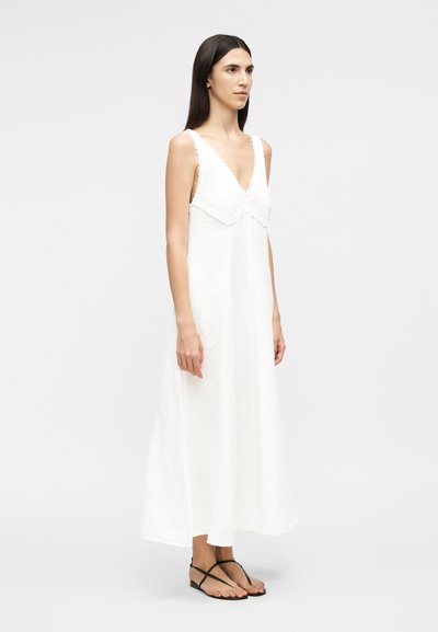 Hosszú sötét hajú nő ujjatlan fehér maxi ruhában, melynek horgolt V-nyakú felső része van, és fekete pántos lapos szandált visel, oldalról áll.