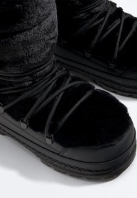 Bottes d'hiver noires à texture de fourrure, semelles épaisses en caoutchouc et lacets croisés, portées avec un pantalon noir duveteux.