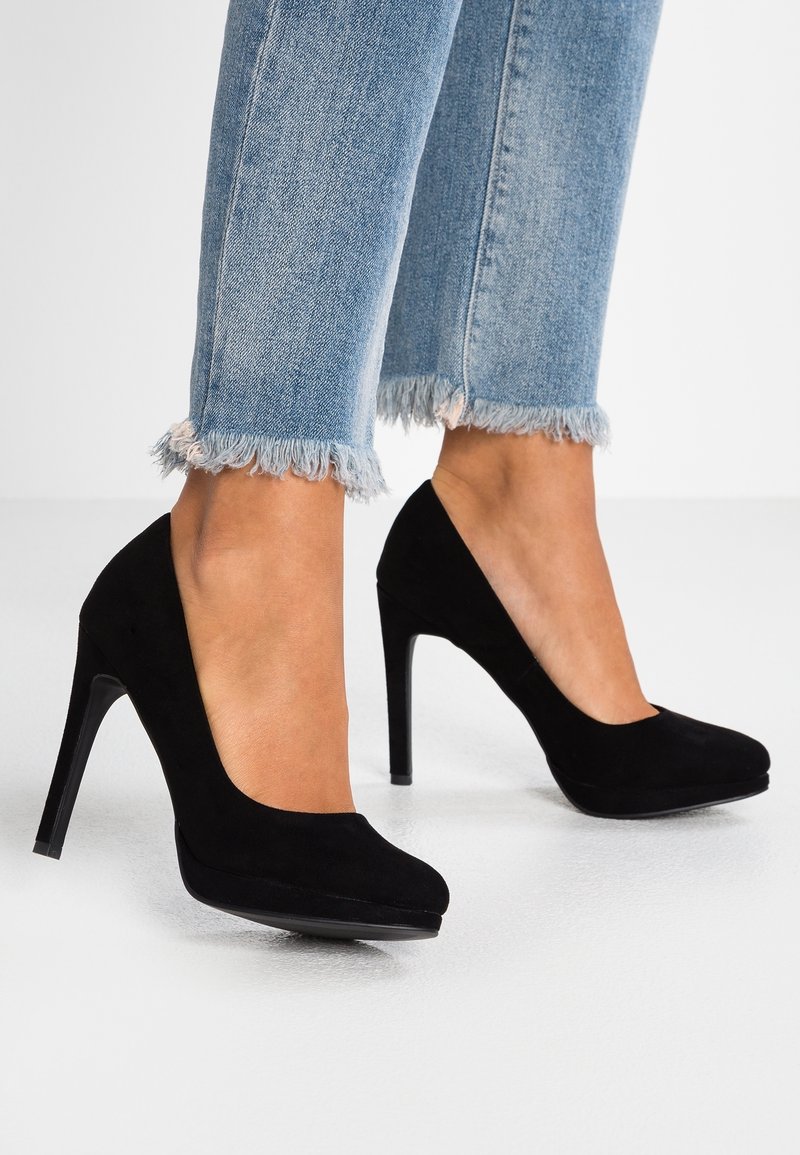 Femme portant un jean bleu clair à ourlet effiloché et des escarpins noirs en daim à talons hauts, debout sur un sol blanc devant un fond blanc.