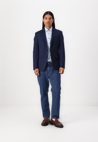 Navy blazer with a notched collar, light blue shirt with buttons, blue jeans, and brown loafers. Smooth fabric and tailored fit.