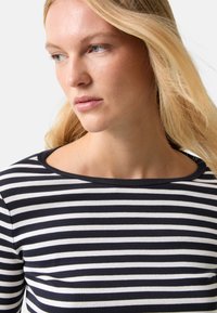 Blonde woman with light skin wearing a black and white horizontal striped top, looking down and to her left against a plain background.