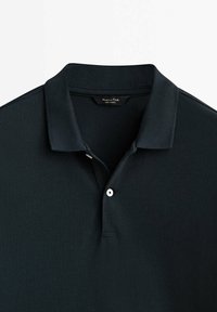 Polo shirt vert foncé en tissu texturé, avec un col classique, une patte de boutonnage à deux boutons et une petite étiquette de marque à l'intérieur du cou.