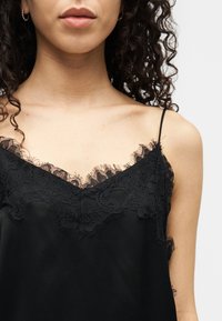 Γυναίκα με σγουρά μαλλιά που φοράει μια μαύρη καμισόλ με δαντελένιο τελείωμα και ένα χρυσό κρίκο στο αυτί, ορατή από τους ώμους έως τα χείλη.