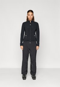 Juoda sportinė striukė su užtrauktuku, ilgomis rankovėmis ir užtraukiamais kišenėmis, derinama su juodomis kelnėmis ir slidinėjimo batais, prieš vientisą foną.